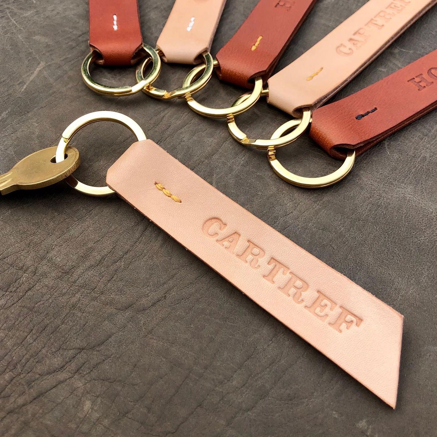 A handmade keyring lies on a grey leather background. The keyring is made from natural coloured leather with yellow stitching and the word "Cartref" stamped into it. It has a flat brass split ring with a key attached to it. There are 5 other leather keyring visible in the background in tan and natural leathers with cream, yellow and black stitching