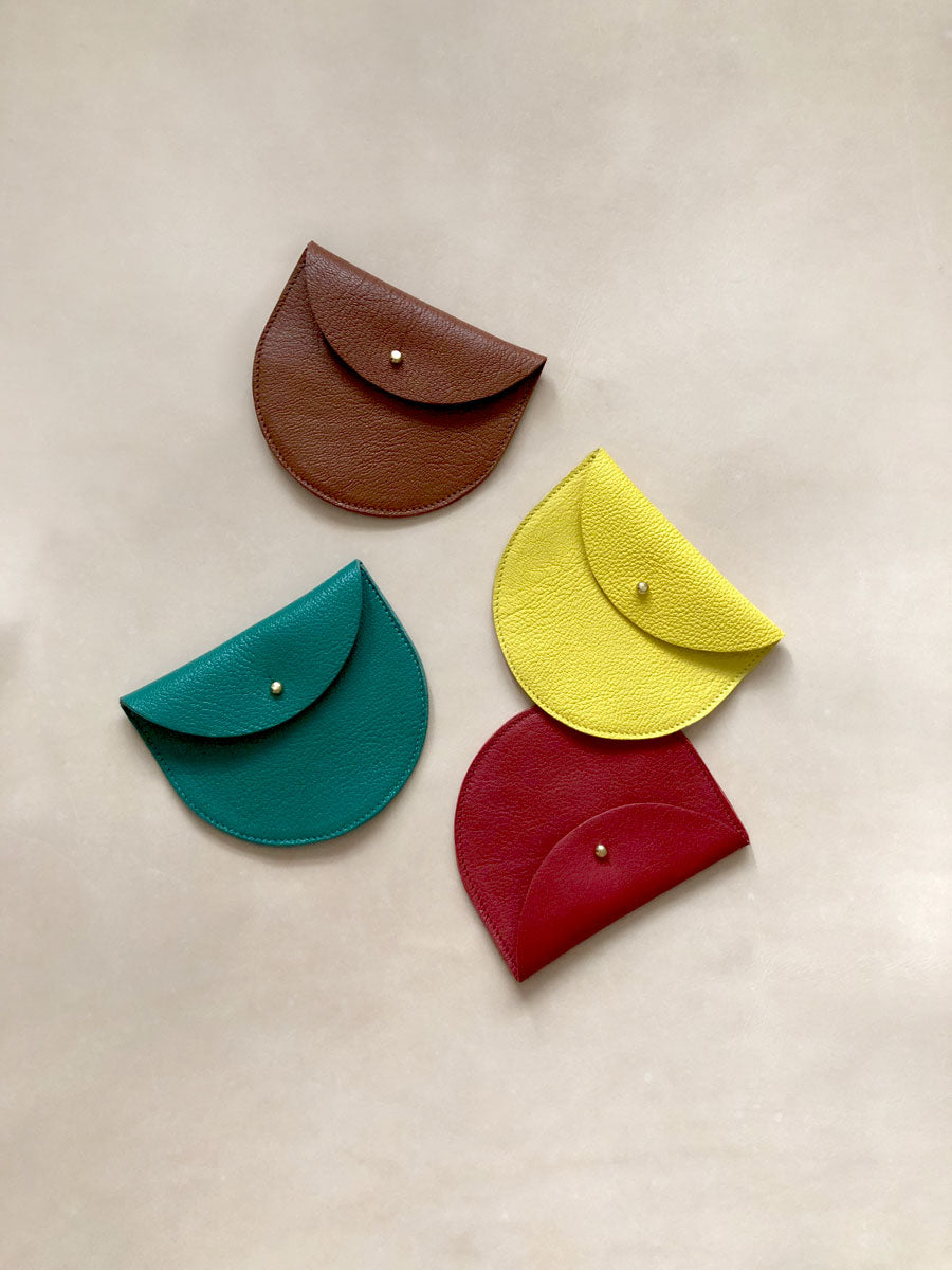 A collection of four simple handmade leather coin purses scattered on a natural leather background. The purses are tan coloured, yellow, green and red.