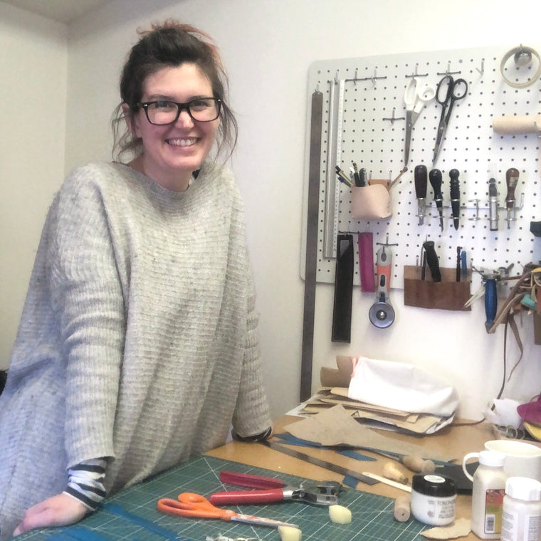 Anna standing behind her workbench in her leatherwork studio. There are various leather working tools scattered on the bench in front of her and hanging on the wall behind her.