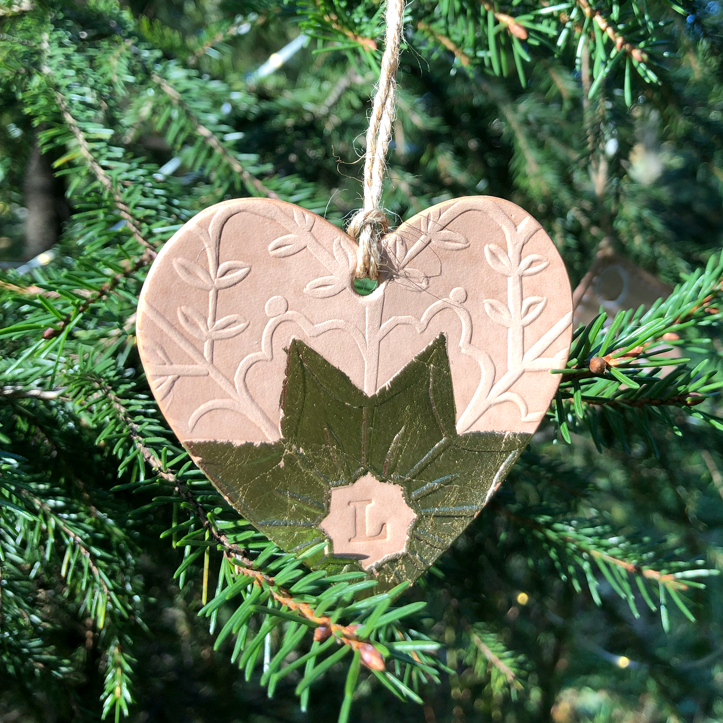 An embossed leather heart shaped Christmas decoration hangs in a tree. It  has been embellished with gold metal leaf a star shape at the bottom and has a letter L stamped into it