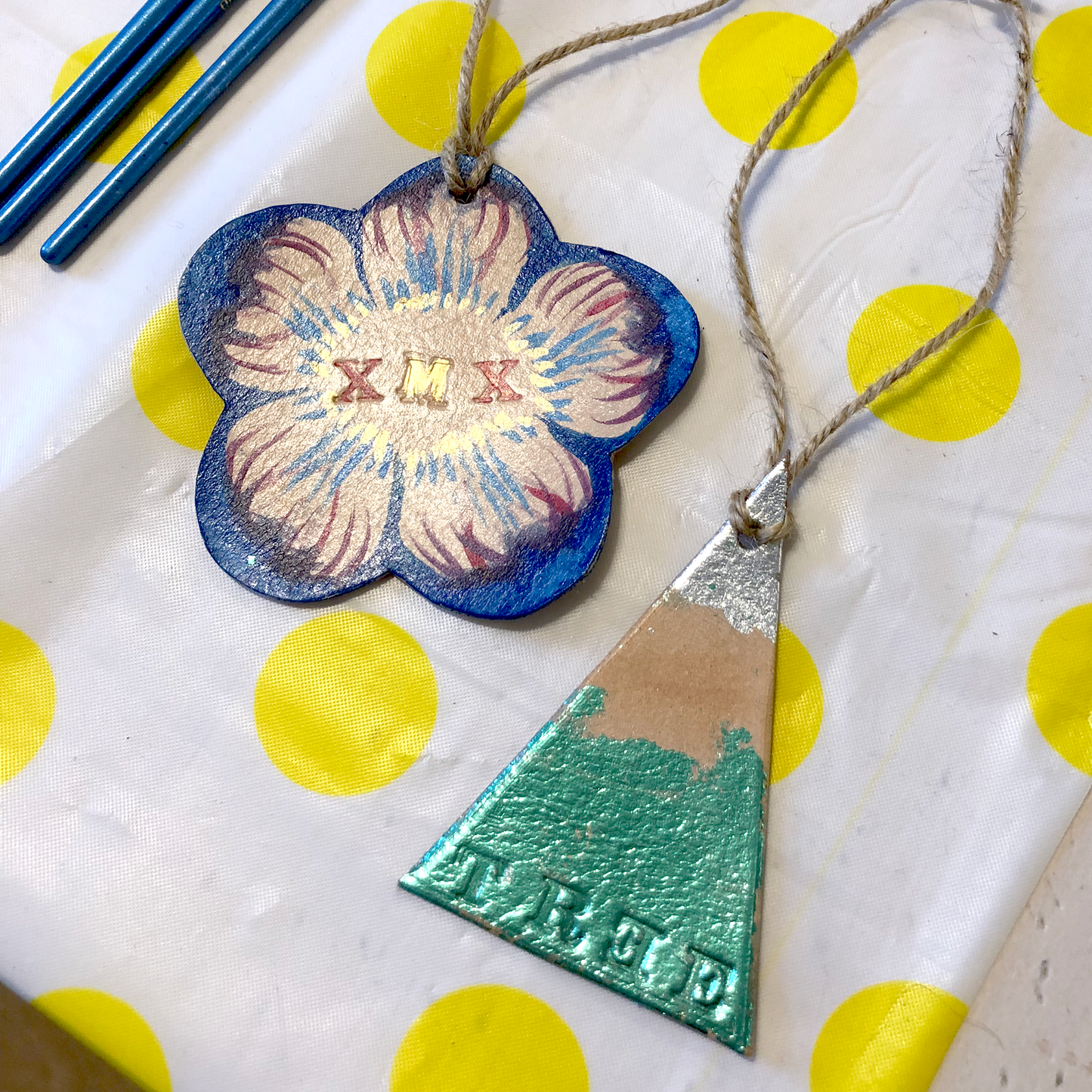 TWO LEATHER CHRISTMAS DECORATIONS LIE ON A SPOTTY BACKGROUND. ONE IS A SIMPLE CHRISTMAS TREE IN THE SHAPE OF A TRIANGLE, WITH SILVER COLOURED METAL LEAF AT THE TOP AND GREEN AT THE BOTTOM. IT HAS THE WORD TREE STAMPED INTO IT. THE OTHER DECORATION IS A FLOWER SHAPE AND HAS BEEN PAINTED IN BLUE, RED AND GOLD