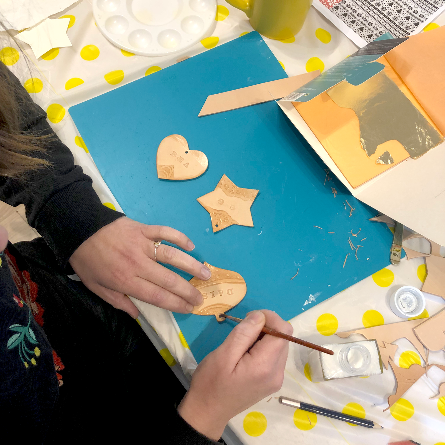 A PAIR OF HANDS IS USING A BRUSH TO APPLY SIZE TO A LEATHER CHRISTMAS ORNAMENT BEFORE APPLYING METAL LEAF. THERE ARE 2 OTHER DECORATIONS LYING ON THE CUTTING MAT NEXT TO IT