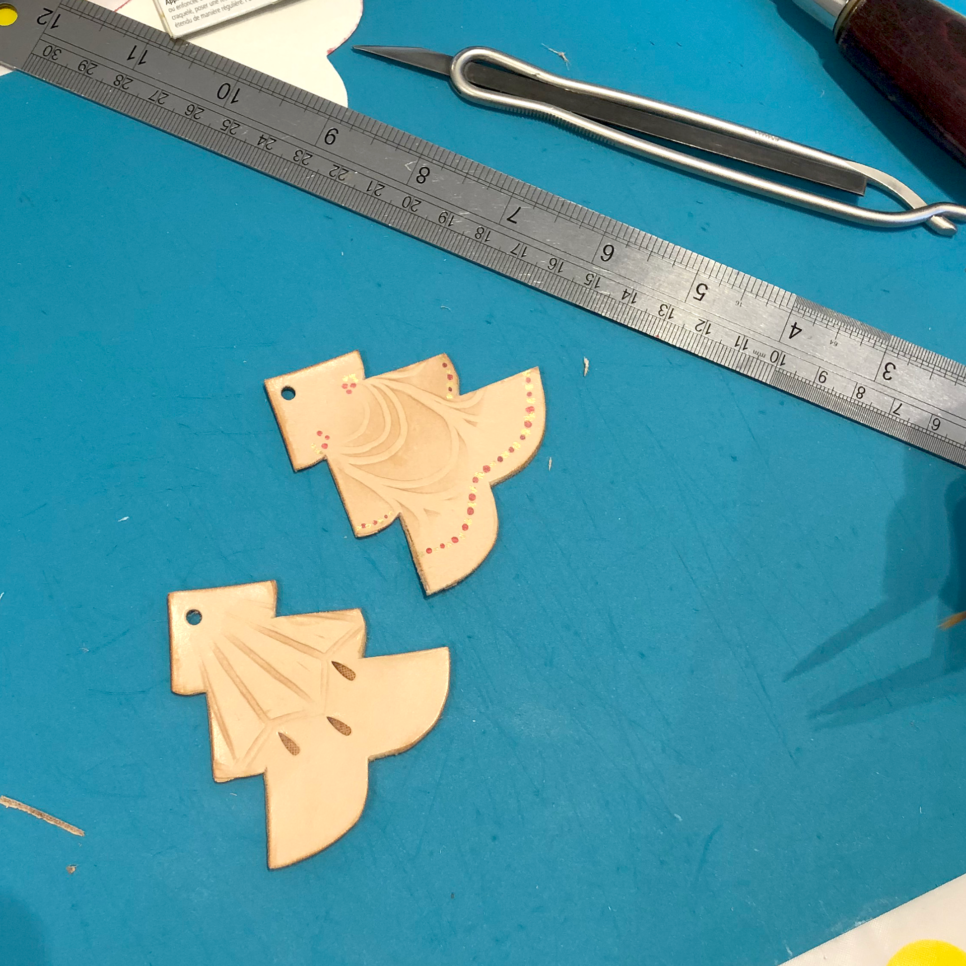 2 CHRISTMAS TREE HAPED LEATHER CHRISTMAS DECORATIONS LIE ON A WORKBENCH BEFORE HAVING THEIR STRING ATTACHED