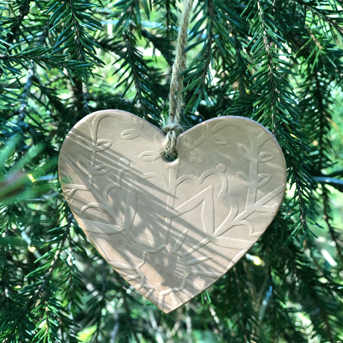 A natural embossed leather heart decoration hangs in a tree. There are shadows of the pine needles visible across it