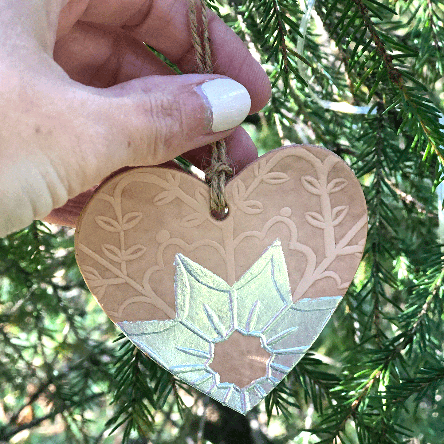 An embossed heart decoration is getting placed in a tree by a hand. The decoration has been embellished with silver metal leaf