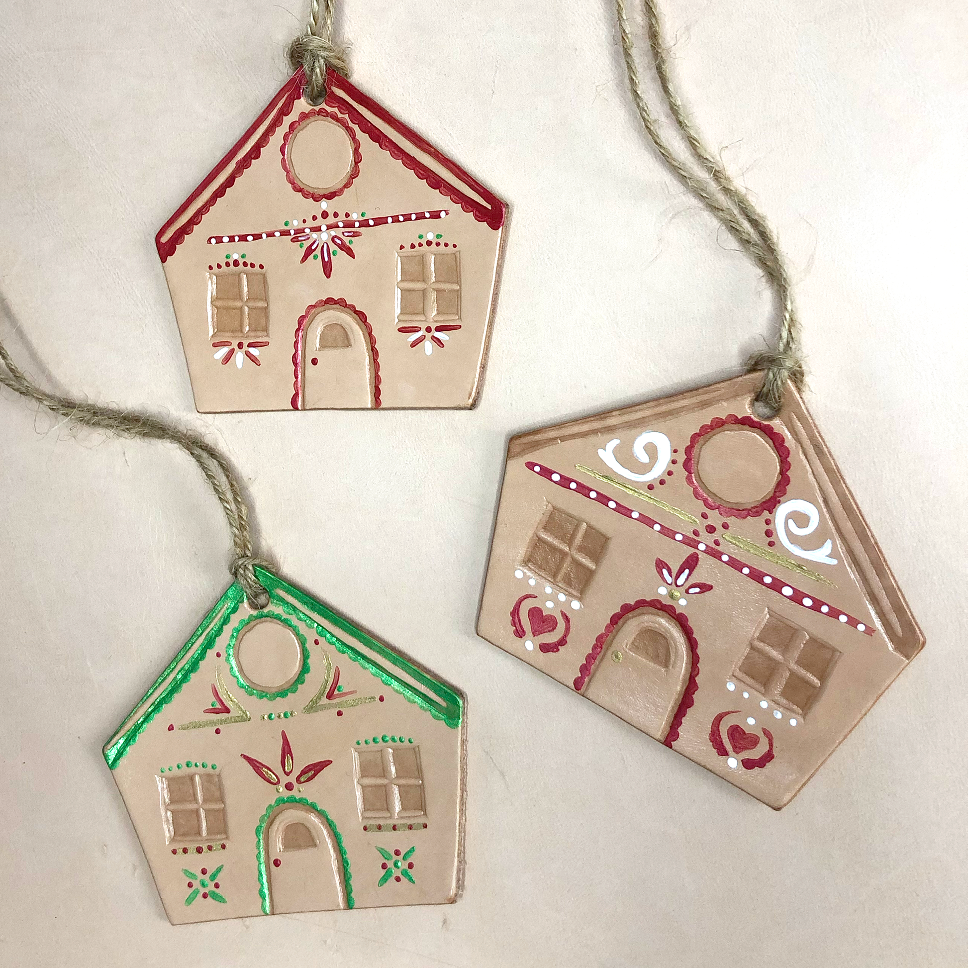 Three hand painted leather gingerbread house shaped Christmas decorations lie on a neutral background. Each one has been painted with a unique design in red, green, gold and white