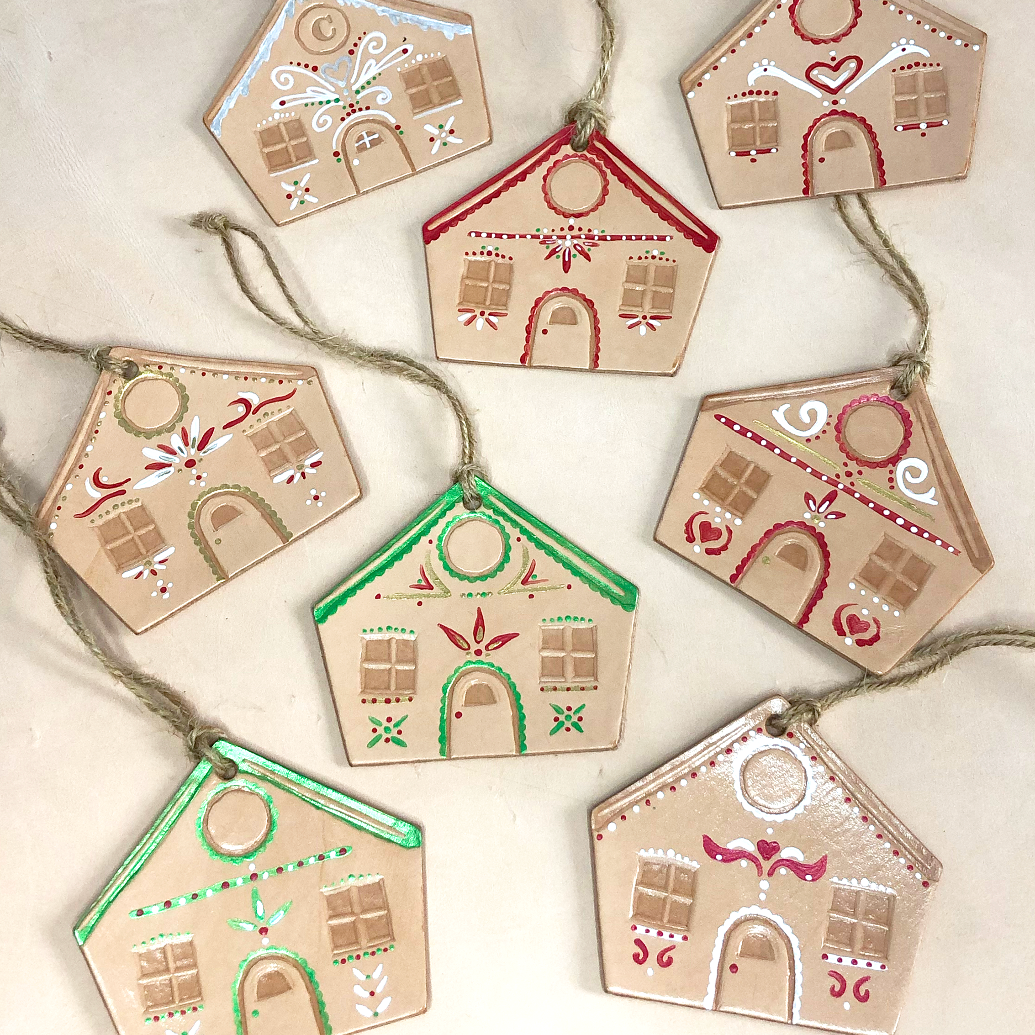 A group of leather gingerbread house decorations lie scattered on a neutral background. Each one has been embossed and painted in a unique festive design use red, gold, green and white paints