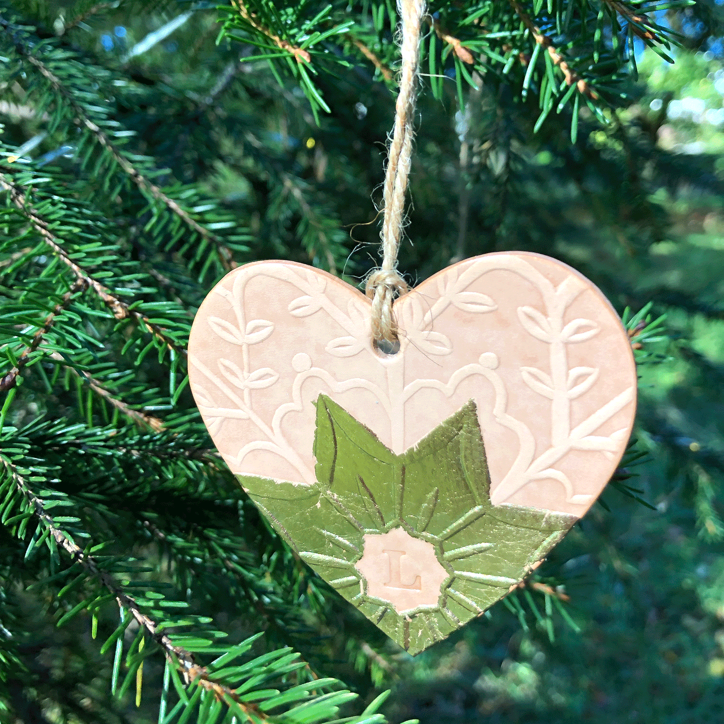 An embossed heart decoration hangs in a tree. It has been embellished with gold metal leaf and has the letter L stamped into it. It is bathed in sunshine