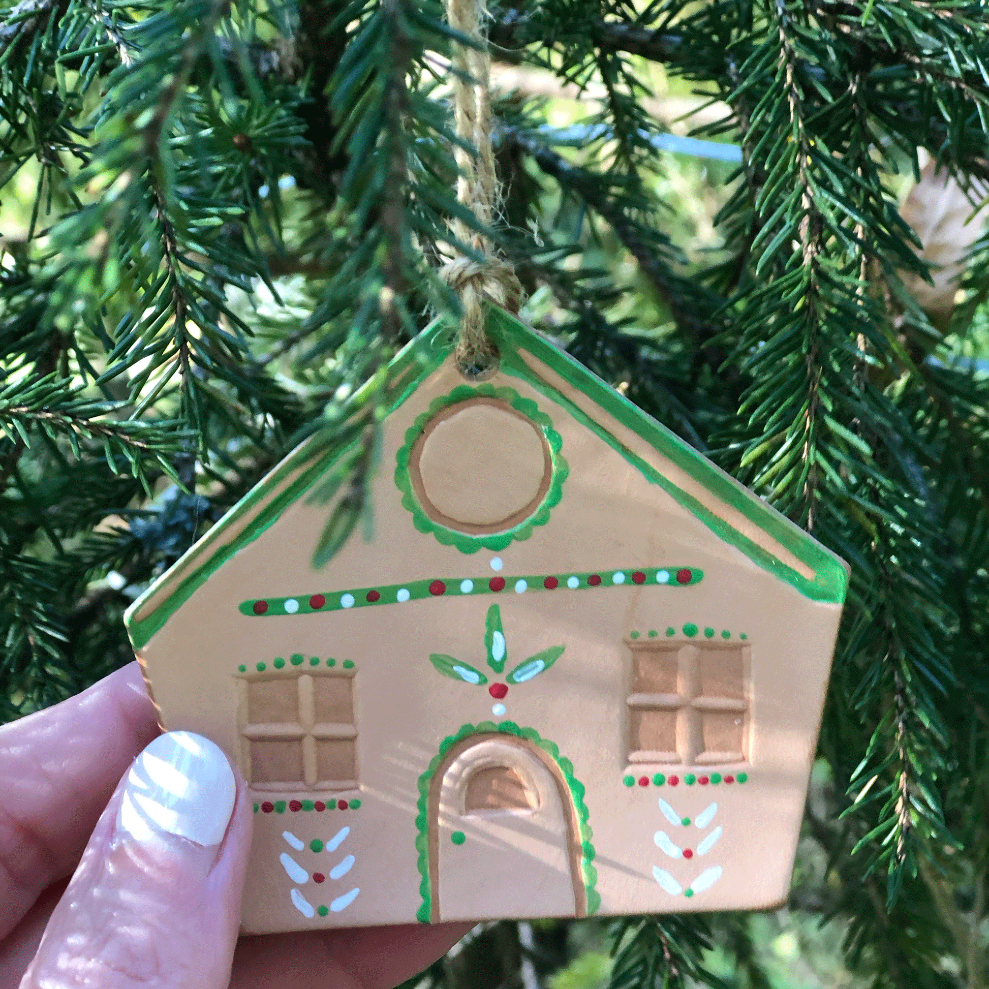 A hand painted leather gingerbread house decoration hangs in a Christmas tree. A hand is touching it. The house has been painted in a festive pattern, mostly in green but with red and white accents