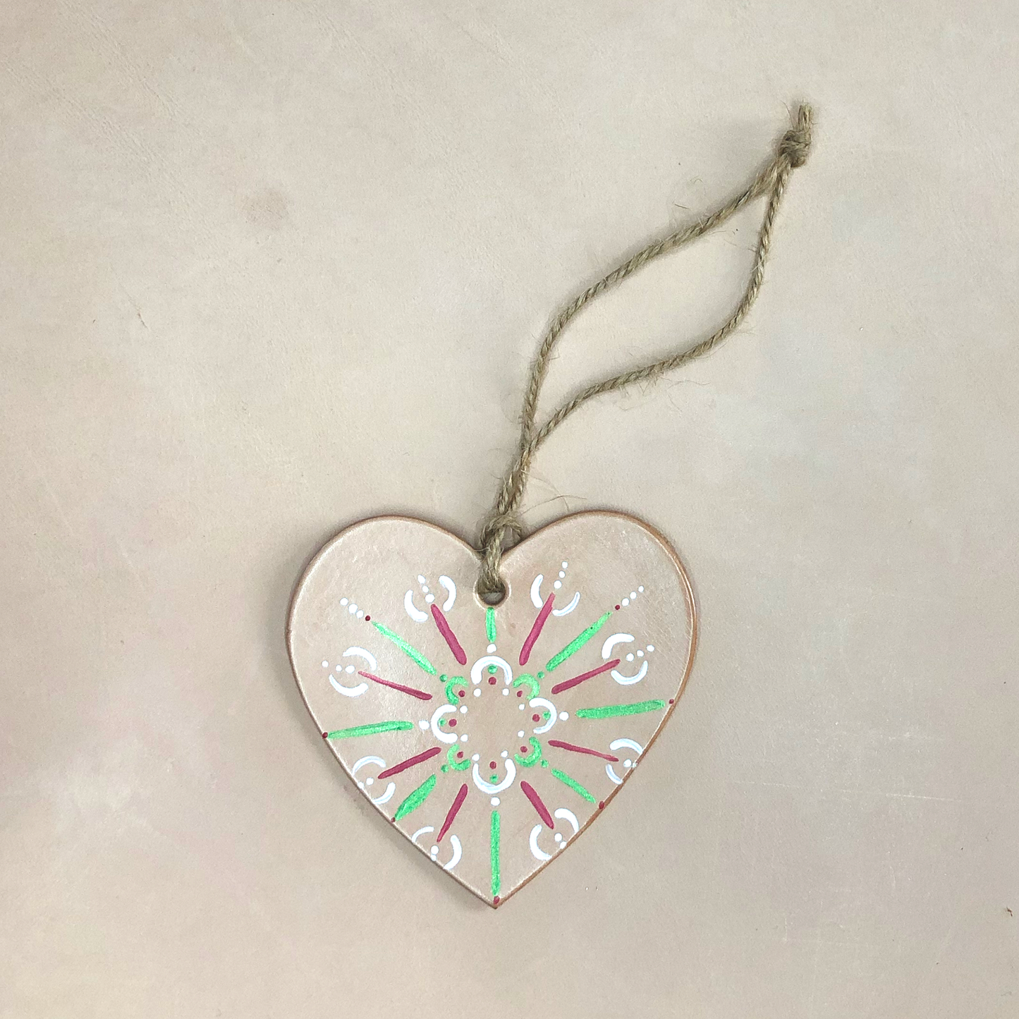 A hand painted heart decoration lies on a neutral background. The decoration is made of natural coloured leather, which has been painted with a red, green and white pattern