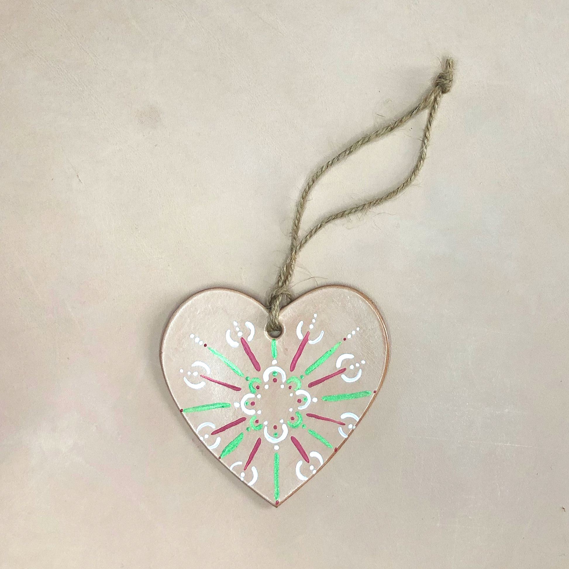 A hand painted heart decoration lies on a neutral background. The decoration is made of natural coloured leather, which has been painted with a red, green and white pattern