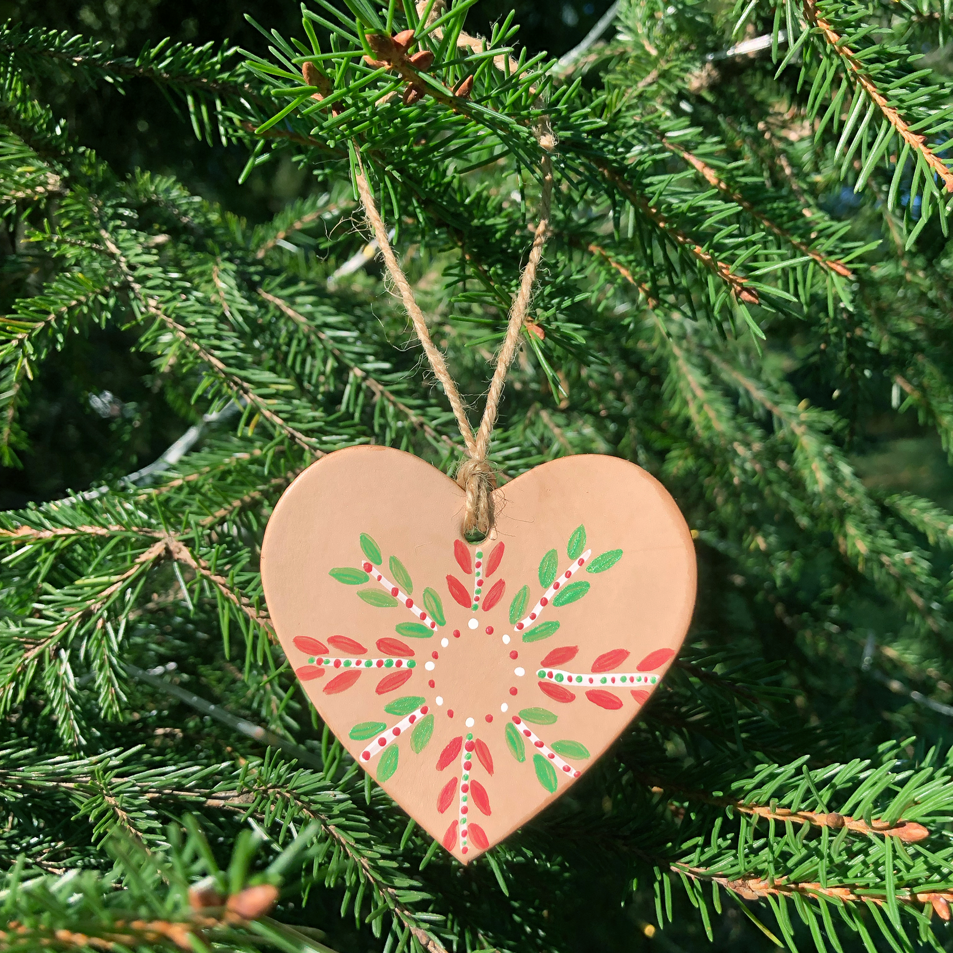 A hand painted leather heart Christmas decoration hangs in a Christmas tree. It has been painted with a festive pattern in red, green and white