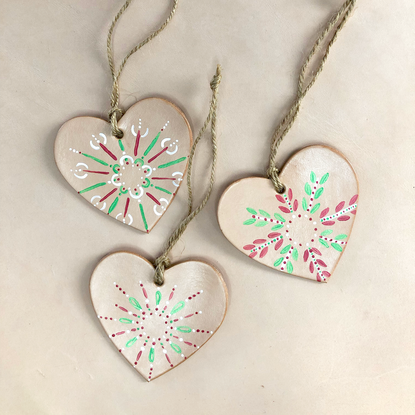 A set of 3 painted leather heart decorations lie on a neutral background. Each heart has been painted with a different pattern in red green and white