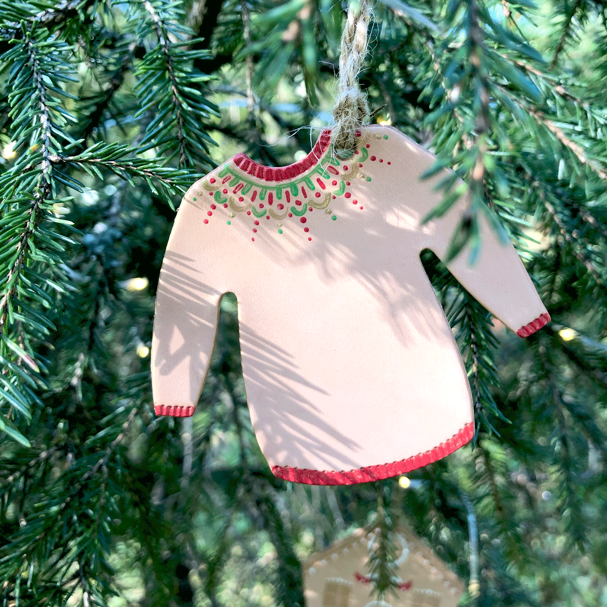 A hand painted leather Christmas jumper decoration hangs in a Christmas tree. Its collar, cuffs and hem have been painted in red and it has a delicate festive design painted around the neckline in red, green and gold.