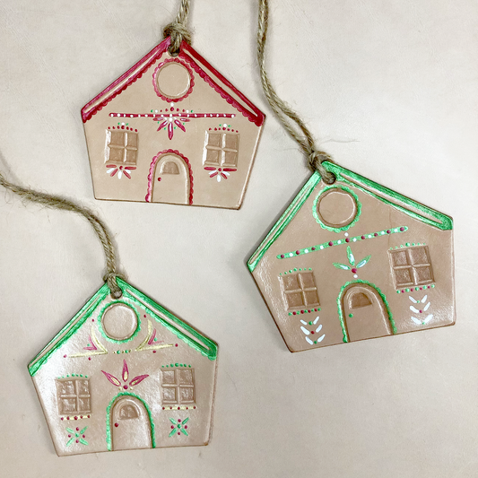 A collection of 3 leather gingerbread house decorations lie on a neutral background. Each has been embossed and painted by hand in a unique design, using green, red, gold and white paints.