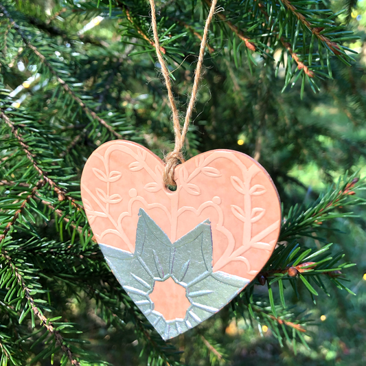 An embossed heart shaped Christmas decoration hangs in a tree. It has been embellished with silver coloured metal leaf.