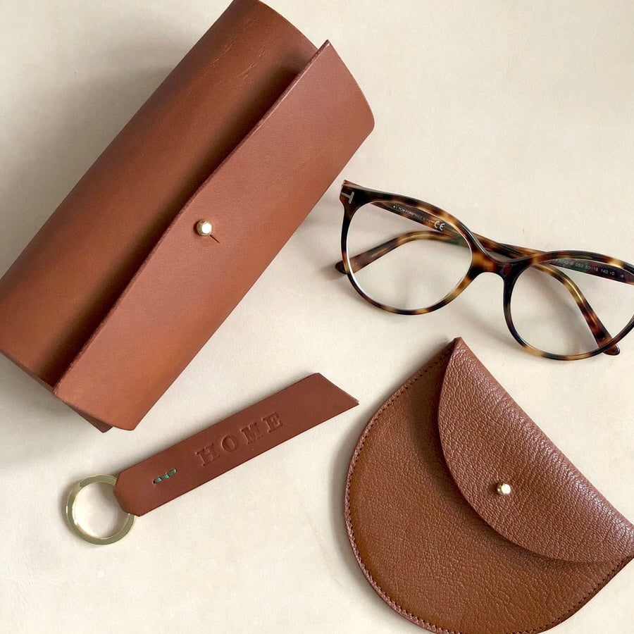 A selection of handmade leather goods photographed from above, alongside a pair of tortoiseshell glasses. There is a glasses case, keyring and purse. All are a traditional tan colour