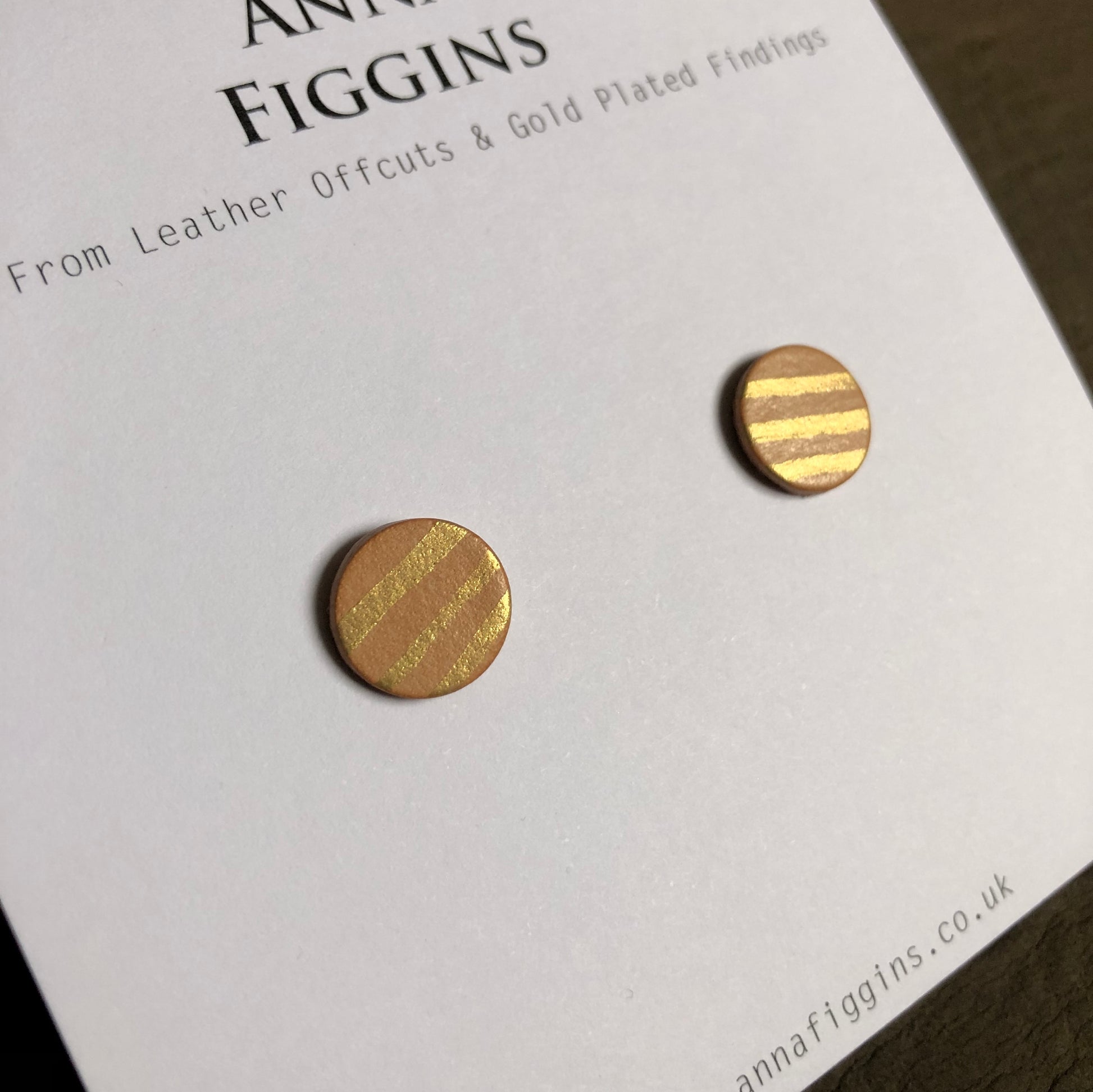 A close up of a pair of leather stud earrings on their asking card. The earrings have been painted by hand in a gold stripe design