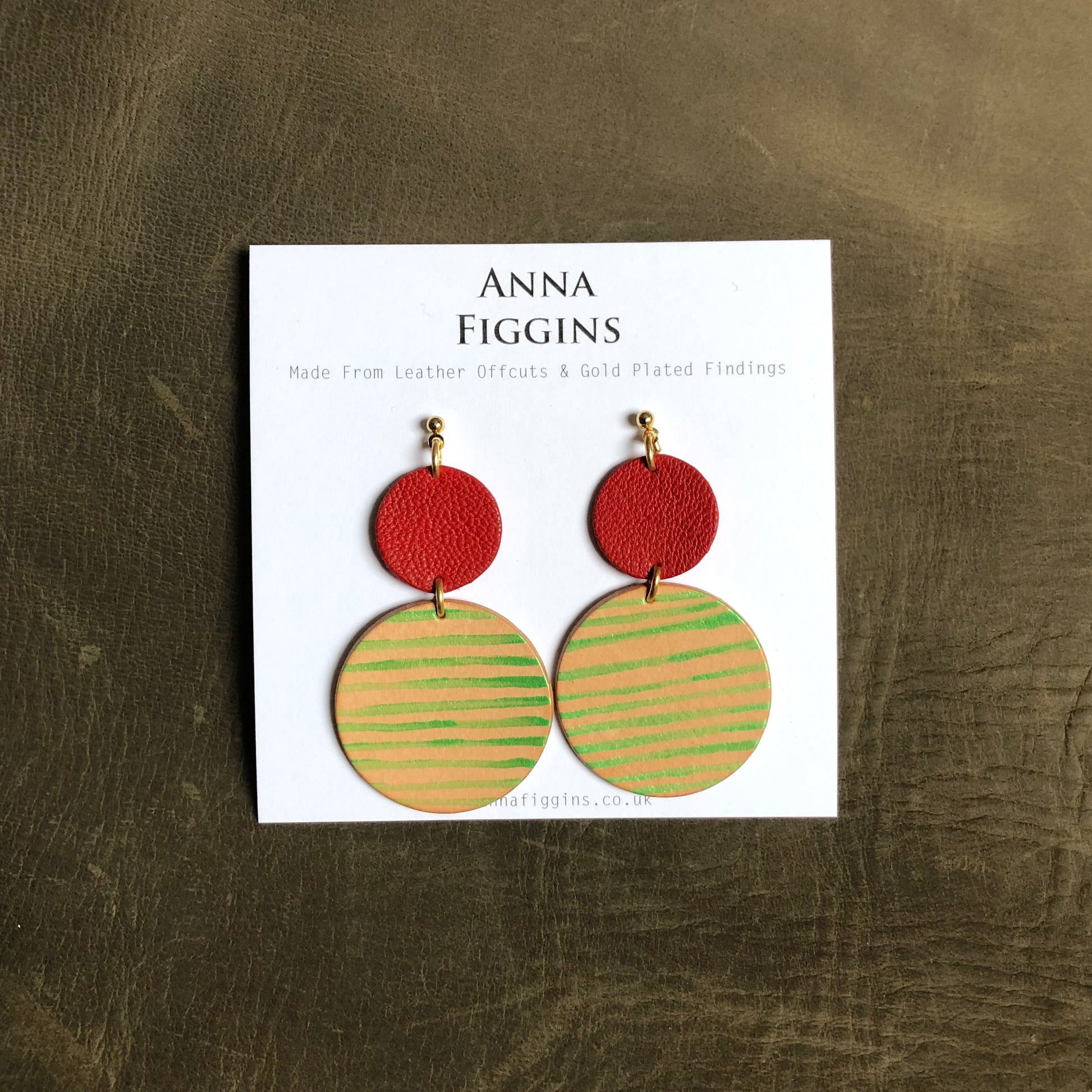 A pair of hand painted leather earrings on their display card. The earrings are Ade from a smaller red leather disc and a larger natural leather disc that has been painted with a metallic green stripe