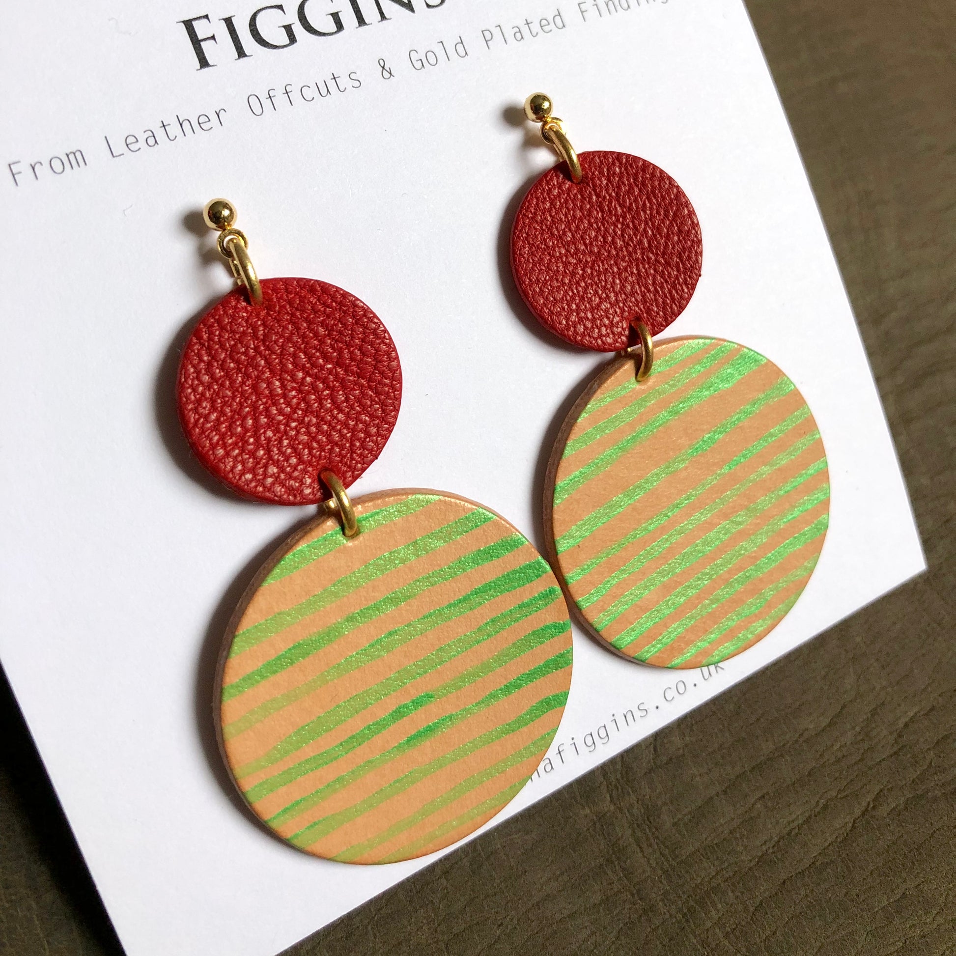 Close up image of a pair of hand painted leather earrings on their display card. They are made of 2 circles, one larger than the other. The smaller circle at the top is a textured red leather, and the larger circle underneath is a natural coloured leather with metallic stripe pattern