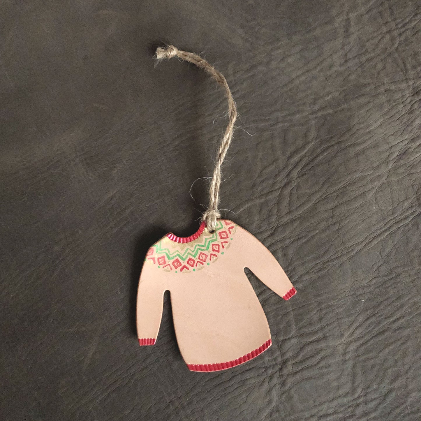 A hand painted leather Christmas jumper decoration lies on a grey leather background. The decoration has been painted with red, green an gold paints in a festive pattern