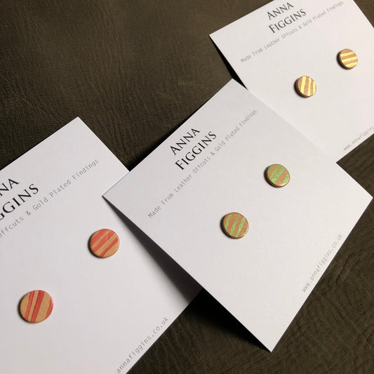 3 pairs of hand painted leather studs on their display cards. The earrings have all been made from natural coloured leather that has been painted with pearlescent paints in a classic stripe pattern. One pair has red stripes, the next green, and the last pair gold stripes.