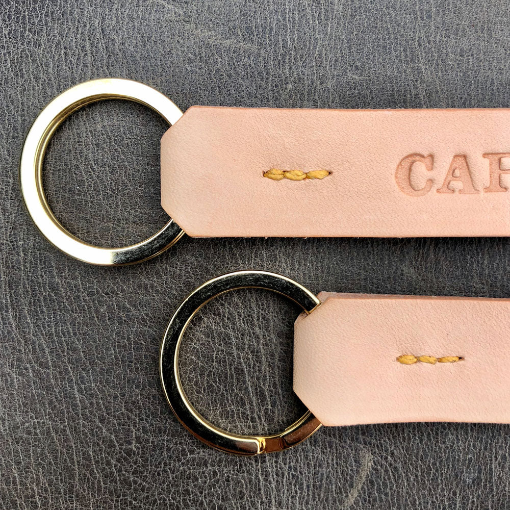 Detail image of the ring ends of 2 keyring. The keyring are made from un-dyed leather and have been stitched with yellow thread. The split rings are brass and you can see some of the letters that have been stamped into the leather of one of the keyrings