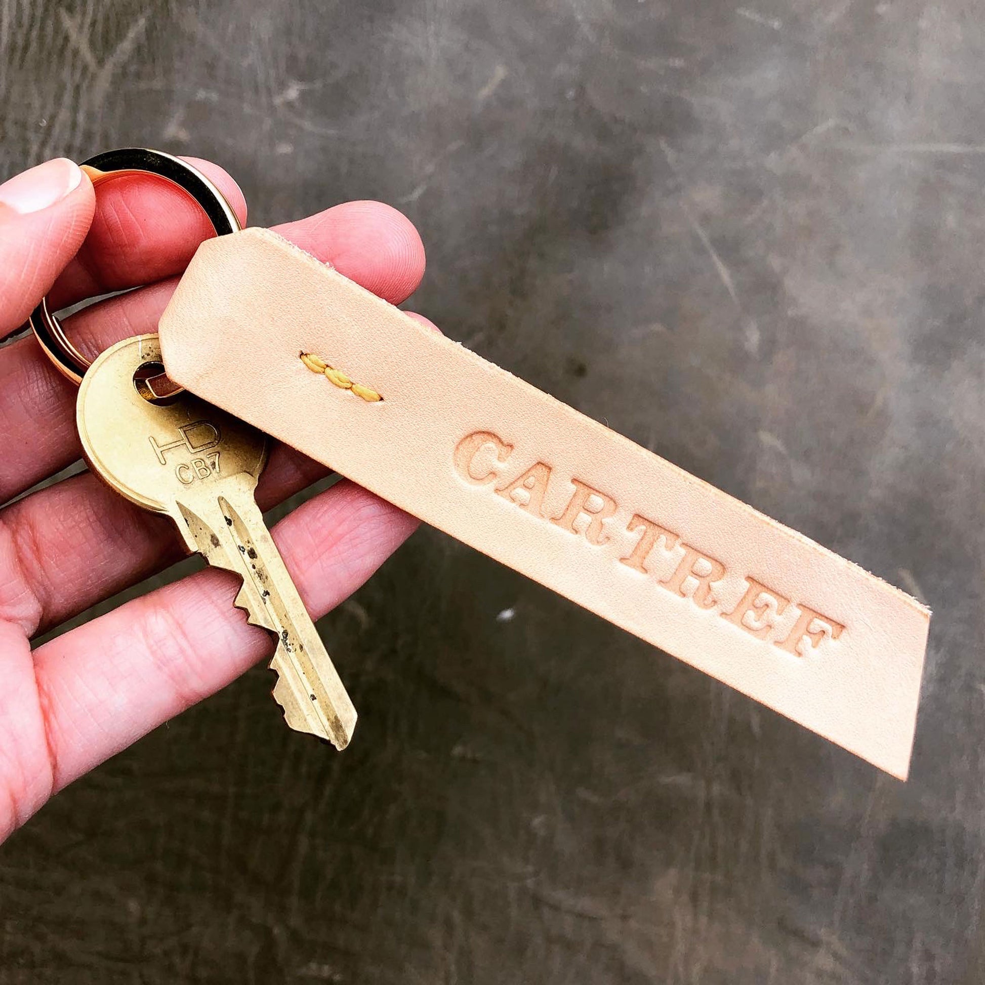 A keyring made from undyed vegetable tanned leather is held in a hand in front of a grey leather background. The keyring has yellow stitching and the word "CARTREF" stamped into it